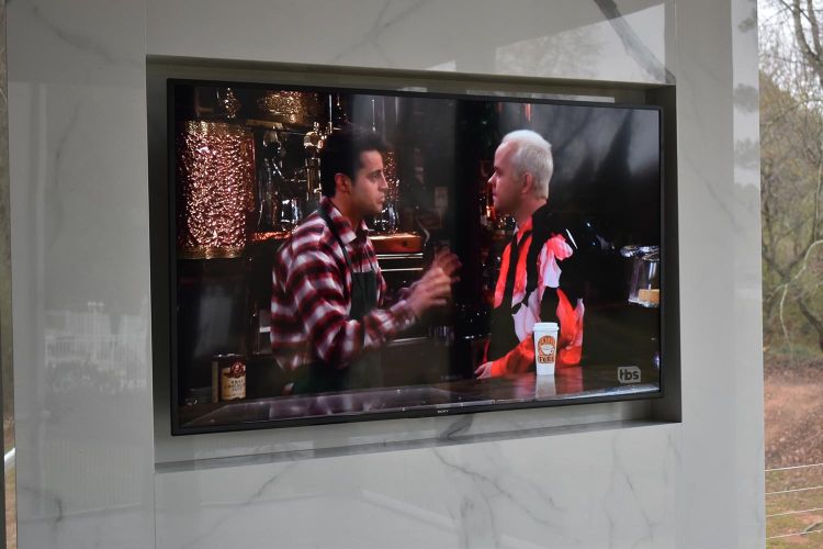 TV embedded in marble wall showing a sitcom scene with two characters at a bar